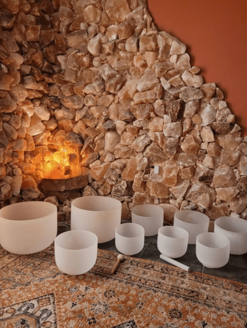 A serene room with a stone wall, featuring various crystal singing bowls arranged on a patterned rug.